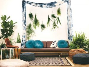 Interior boho, foto de um sofá com almofadas azuis redondas, plantas em ambos os lados e um tecido drapeado que domina a decoração.