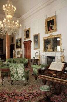 以摄政时期风格装饰的房间。房间中央有一盏华丽的枝形吊灯，还有各种镶着金框的肖像画、绿色碎花家具、一座白色壁炉和一架中木质钢琴。
