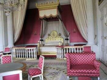路易十四的卧室，有粉红色的圆点家具和一张带有金色装饰的白色大床。