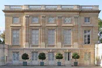 Le Petit Trianon:浅棕褐色的建筑，有巨大的矩形窗户，四棵树沿着前面，五组双层门沿着地面。
