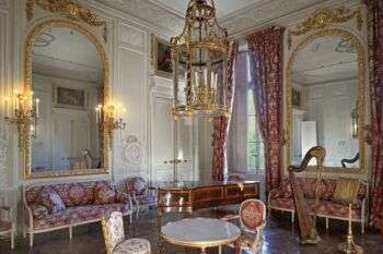 Le salon de compagnie:有红白家具和窗帘的大房间。房间里有两面巨大的拱形金框镜子，中央有一盏金色的枝形吊灯，右边角落有一架竖琴。