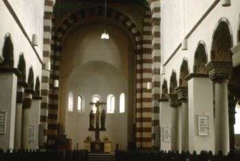 内部设计:一个简单的教堂，有条纹拱门，空白的棕褐色墙壁，在岛的尽头有一个十字架。小拱形窗户位于靠近照片背面的圆顶中间。