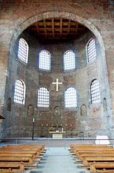君士坦丁大教堂内部的照片。一个圆形的圆顶结构延伸到一个矩形的房间。照片的右下方和左侧是教堂的长凳，中间是一个小岛。此外，还有一个祭坛和岛的尽头和一个白色的十字架坐在祭坛上面。